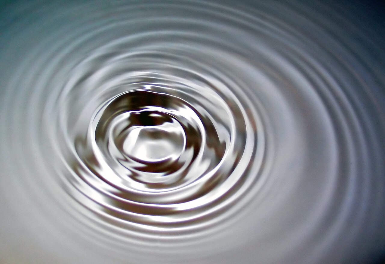 Close-up of a silvery water surface with a small central dip and rings of ripples spreading outward in overlapping circles, against a soft gray-blue background.