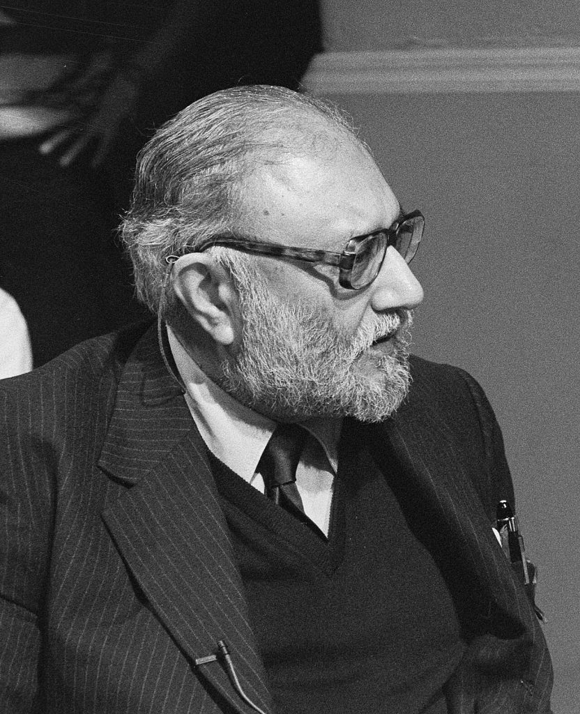 Black-and-white photo of an older man with a gray beard and large glasses, wearing a pinstriped suit, sweater and tie, looking to the right with a thin earpiece cord over his ear.