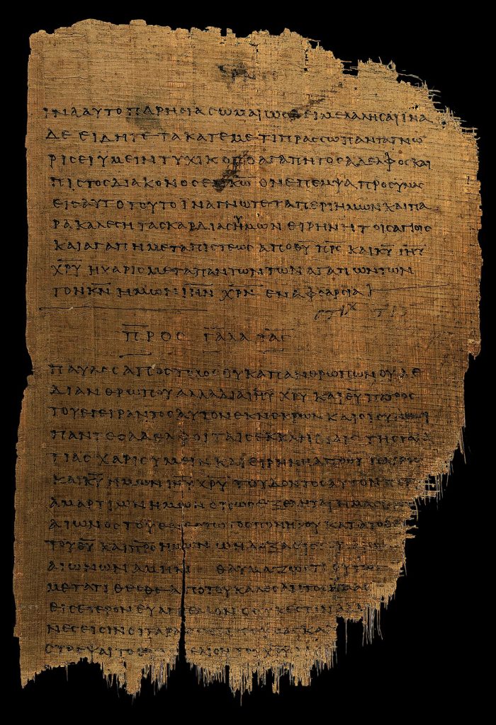 A ragged, tan sheet of ancient papyrus on a black background, covered in rows of handwritten Greek capital letters. Near the middle, a short centered heading, the Greek title of the letter to the Galatians, separates the end of one text from the start of the next. The bottom edge is frayed and split.