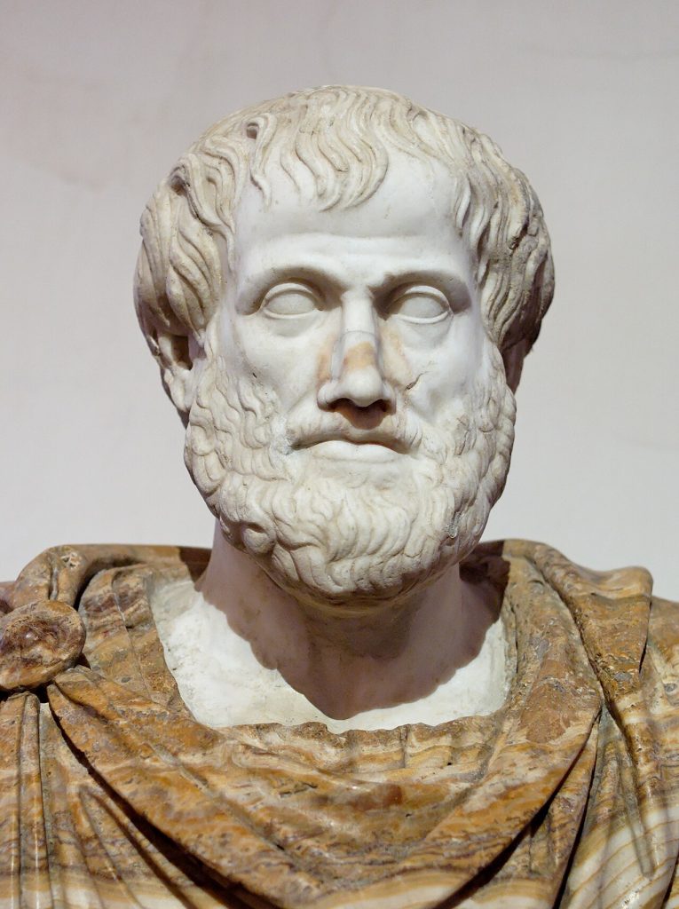 Marble bust of a bearded man with thick wavy hair and a calm face, wearing a draped mantle of banded tan and cream stone, against a plain pale wall.