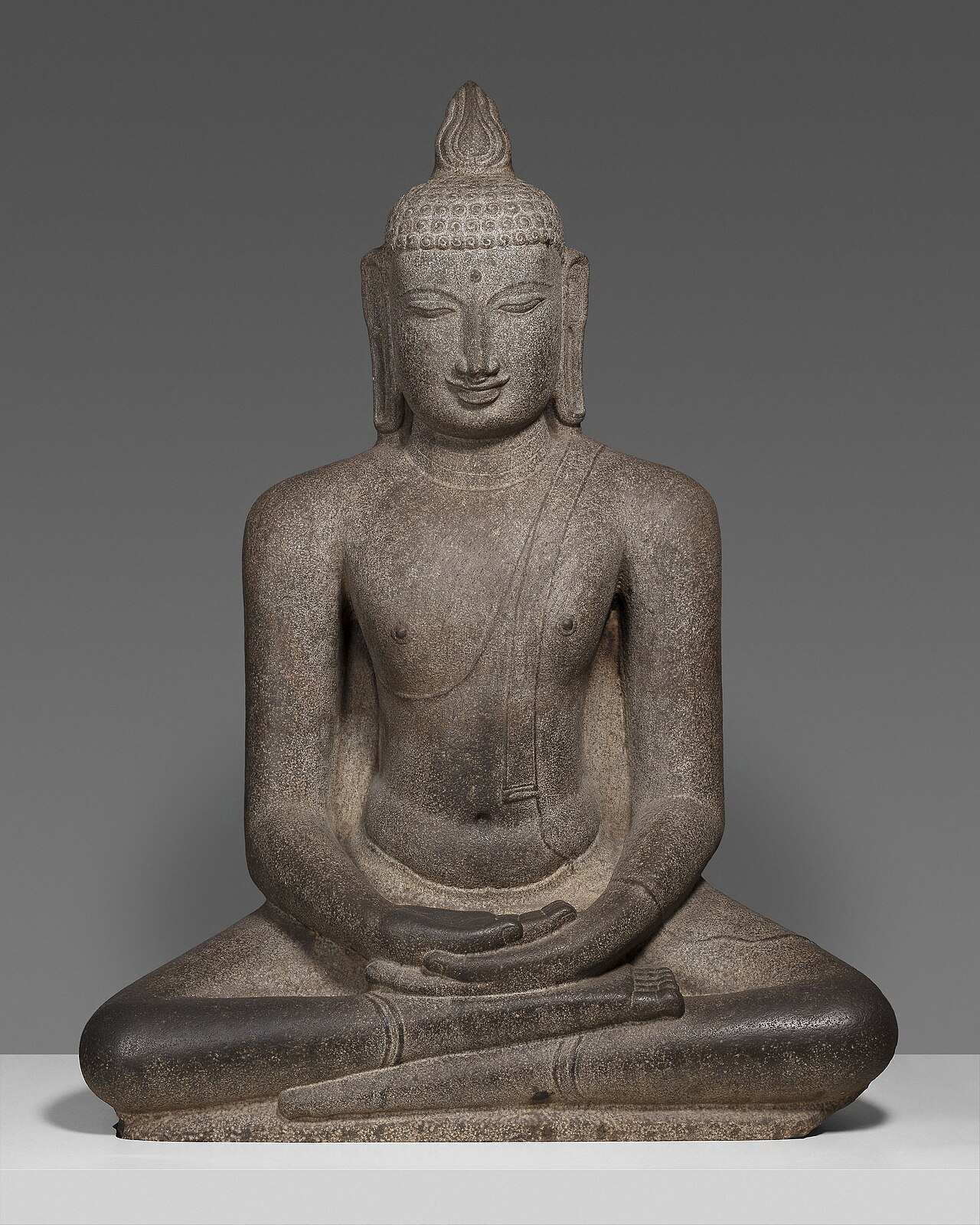 A gray granite statue of the Buddha seated cross-legged, hands resting one on the other in his lap, eyes lowered and a slight smile, with a small flame shape on top of his head, against a plain gray background.