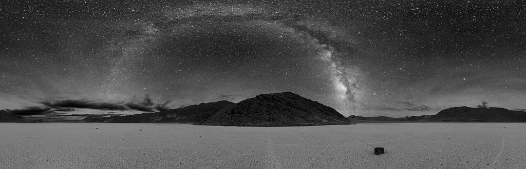 Black-and-white 360-degree panorama of a flat, cracked dry lakebed at night under a sky full of stars. The bright band of the Milky Way arches over a dark rocky hill in the center, and a single rock sits on the lakebed at the lower right.