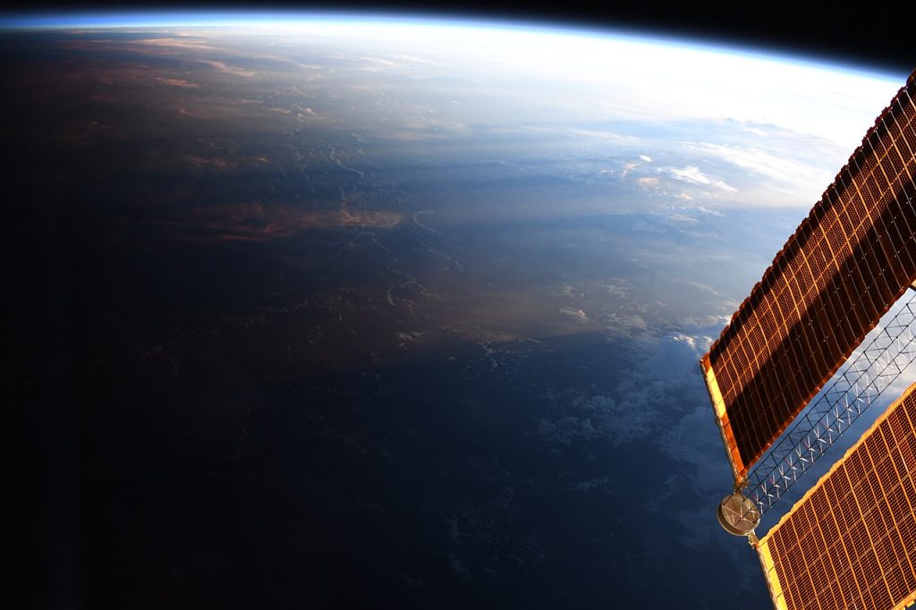 View from the International Space Station of Earth's curved surface, split between light and dark. A bright blue horizon and sunlit clouds at the upper right fade into deep shadow at the lower left, with golden solar panels along the right edge.