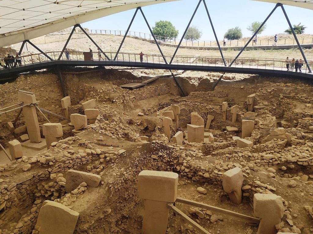 An excavated site of low stone walls and tall T-shaped stone pillars in pale earth, seen from above, some pillars braced with wooden supports, with a curved walkway, a large white canopy on steel beams overhead, and visitors standing along the railing.