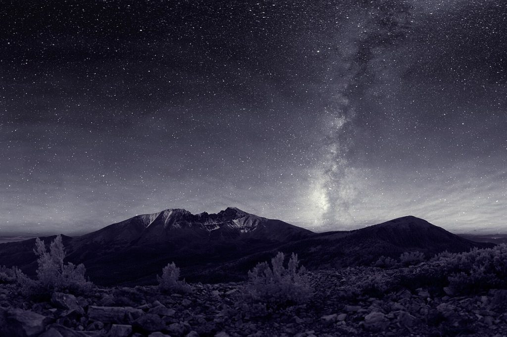 Black-and-white night photo of the Milky Way rising in a bright band over a snow-streaked mountain, with a sky full of stars above a rocky slope and small shrubs.