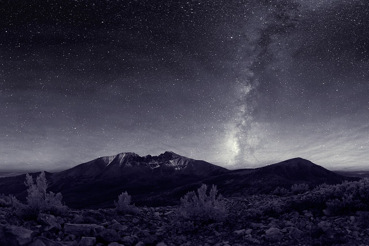 Black-and-white night photo of the Milky Way rising in a bright band over a snow-streaked mountain, with a sky full of stars above a rocky slope and small shrubs.