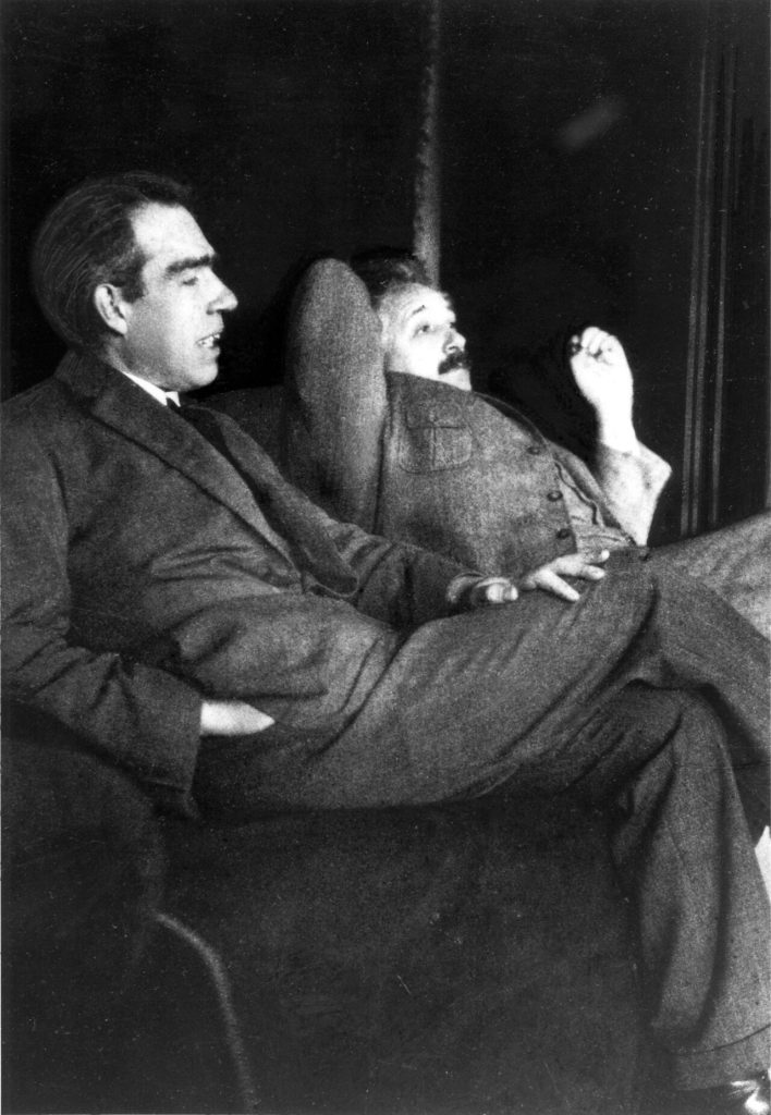 Black-and-white photo of Niels Bohr, seated in the foreground in a dark suit and speaking, with Albert Einstein leaning back behind him, one hand behind his head and the other raised.