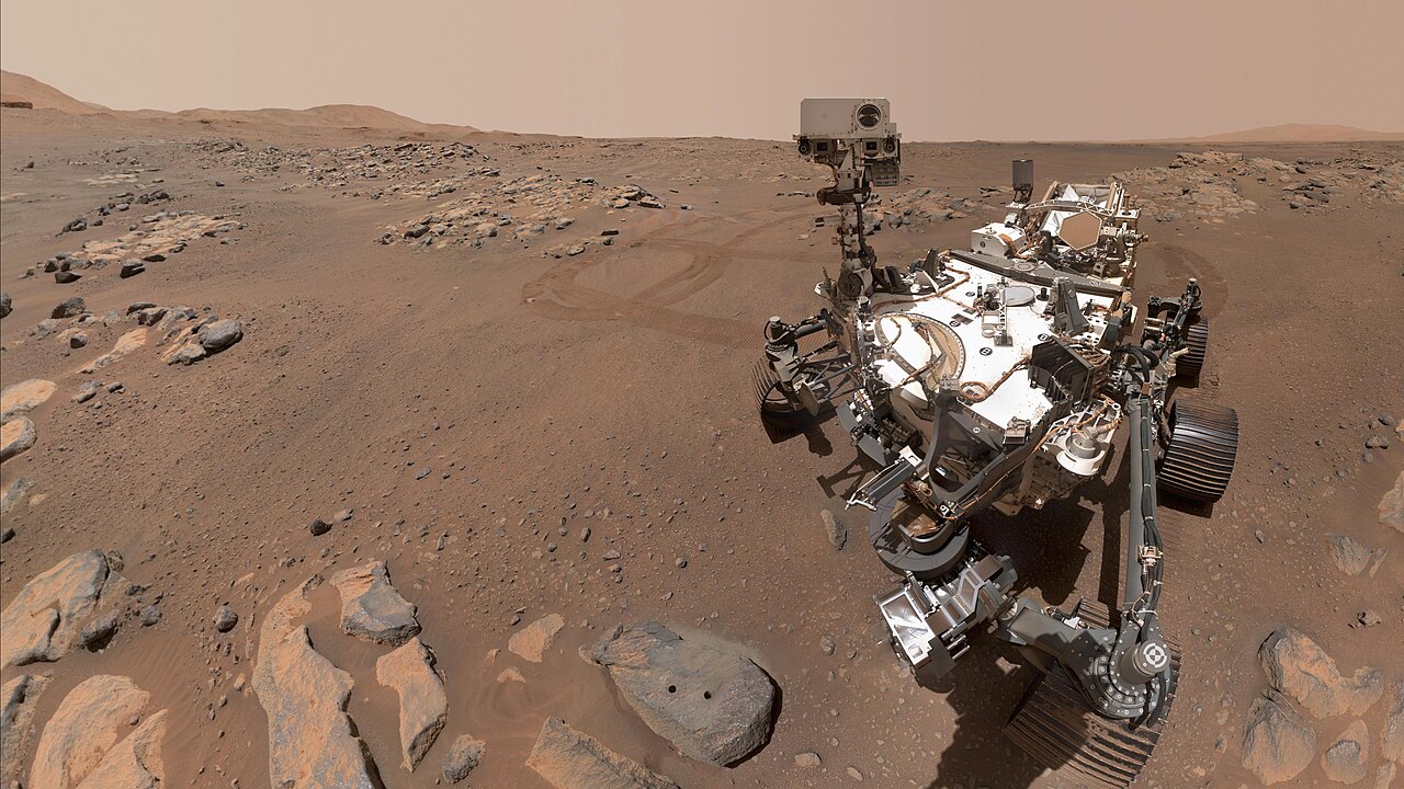 A white six-wheeled rover with a tall camera mast stands on reddish, rocky Martian ground under a hazy tan sky, beside a flat gray rock with two small drilled holes in the foreground.