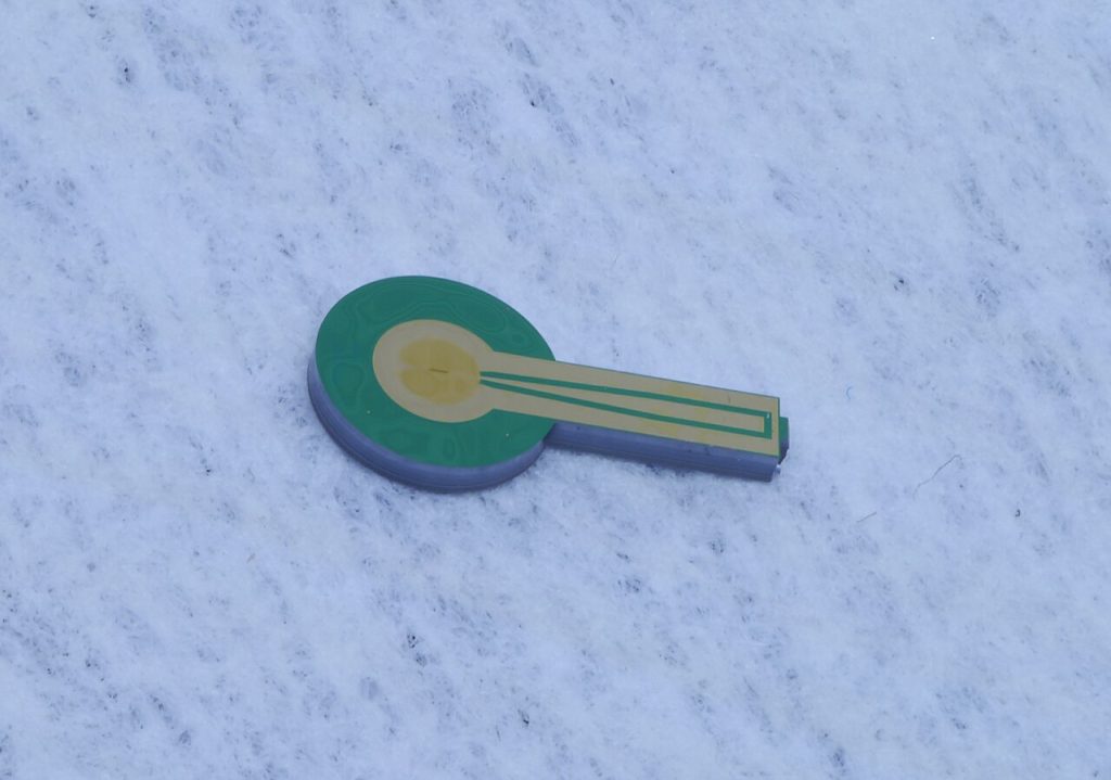 A small key-shaped chip with a round green end, a gold circle at its center and a gold stem, resting on a white, fuzzy surface.