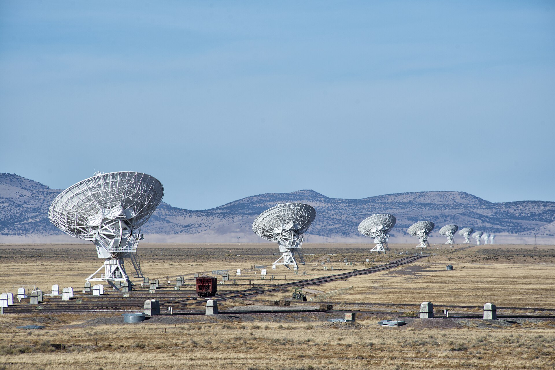 Large white radio dishes stand in a row across a dry, grassy plain, shrinking into the distance toward low brown hills under a pale blue sky. Rail tracks for moving the dishes cross the foreground.