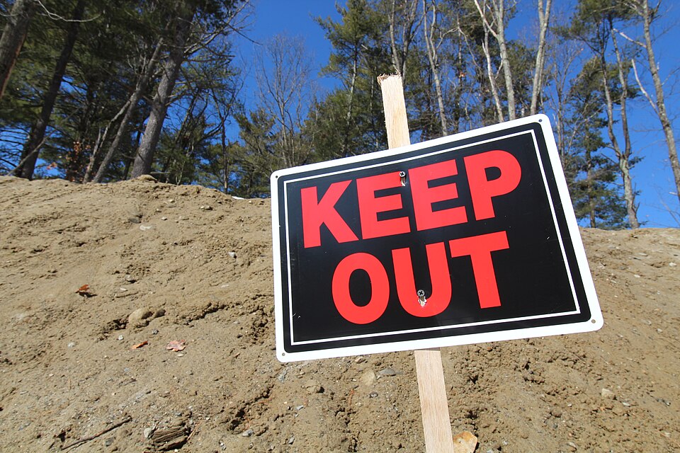 A black sign with large red letters reading KEEP OUT, screwed to a wooden stake on a bare dirt slope, with pine trees and a bright blue sky behind it.