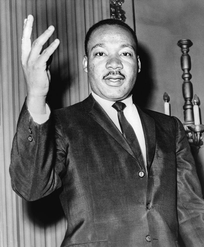 Black-and-white portrait of Martin Luther King Jr. in a suit and tie, one hand raised as he speaks.