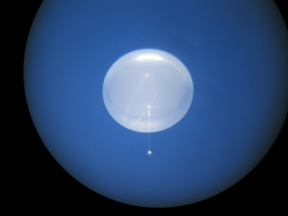 A glowing, pale white dome-shaped balloon floats in a deep blue sky, seen through a round viewfinder, with a thin line hanging below it to a small bright point.