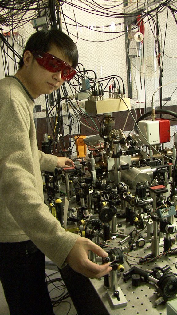 A researcher in a tan sweater and red laser-safety glasses adjusts a small mount on a crowded optical table covered with mirrors, lenses and metal posts, with a cylindrical metal chamber and many cables hanging overhead.