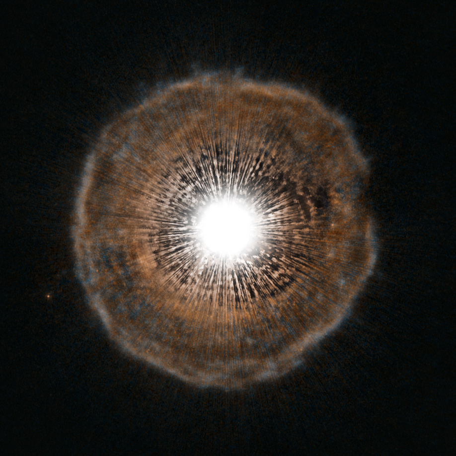A bright white star with streaks of light radiating outward, sitting inside a round, faint orange and blue shell of gas against a black background.