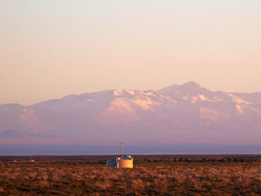 A pale cylindrical water tank topped with an antenna mast stands in dry, reddish scrubland at sunrise. Two more tanks are tiny specks in the distance, and a snow-streaked mountain range glows pink behind.