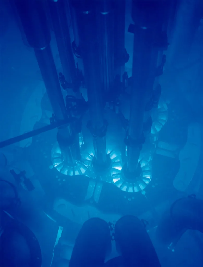 Looking down through deep water into a nuclear research reactor: tall metal tubes and fittings surround a core that glows an intense electric blue.