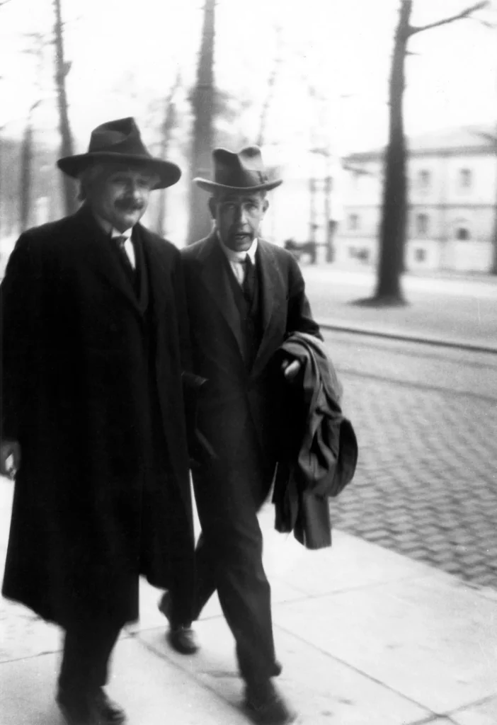 Black-and-white photo of Albert Einstein, left, in a long dark overcoat and hat, walking along a tree-lined street beside Niels Bohr, who wears a suit and hat and carries a coat over his arm.