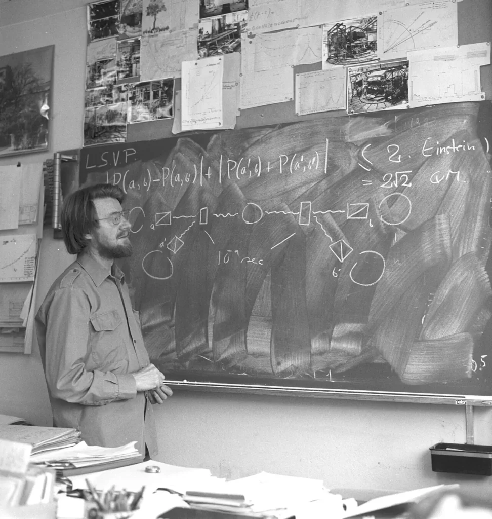 Black-and-white photo of John Bell, a bearded man with glasses, standing beside a chalkboard with a diagram of a two-sided photon experiment and an inequality comparing 2 with 2 times the square root of 2.