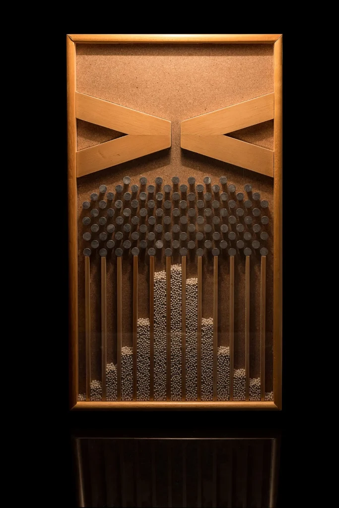 A wooden Galton board in a dark room: rows of dark pegs sit above narrow vertical channels, where small pale beads have piled into columns that are tallest in the middle and shorter toward the sides.
