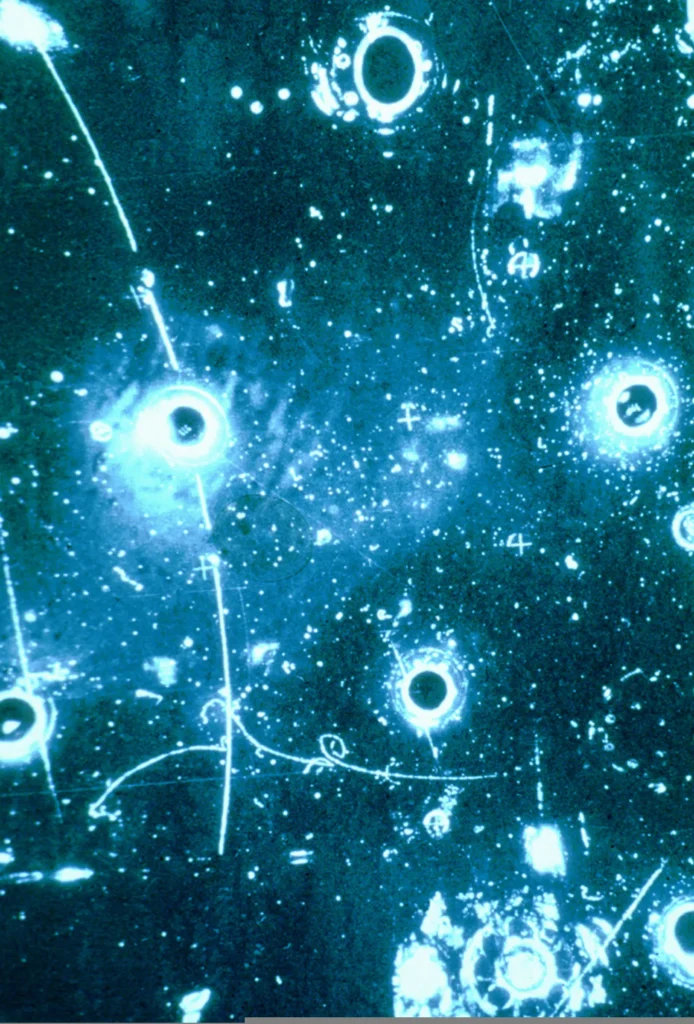 Blue-tinted bubble chamber photograph dotted with bright ring-shaped reflections and speckles. In the lower part, a thin track with small curls runs roughly sideways, left by an electron.