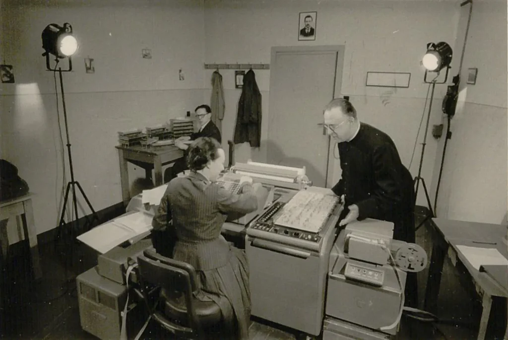 Black-and-white photo of a 1950s computer room. A man in a black clerical cassock and glasses leans over a large desk-sized machine while a seated woman works at its keyboard. Another man sits at a desk in the background between two studio lamps.