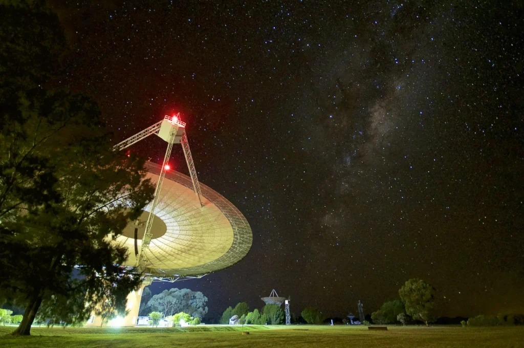 A huge white radio telescope dish, lit from below, tilts toward a starry night sky, with the faint band of the Milky Way across the right side. Trees and a smaller dish stand on the grass below.