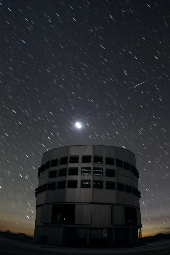 Long-exposure night photograph of a tall, cylindrical telescope building under a sky full of short star trails. A thin streak that brightens in the middle crosses the upper right, and a bright glowing spot hangs above the building.