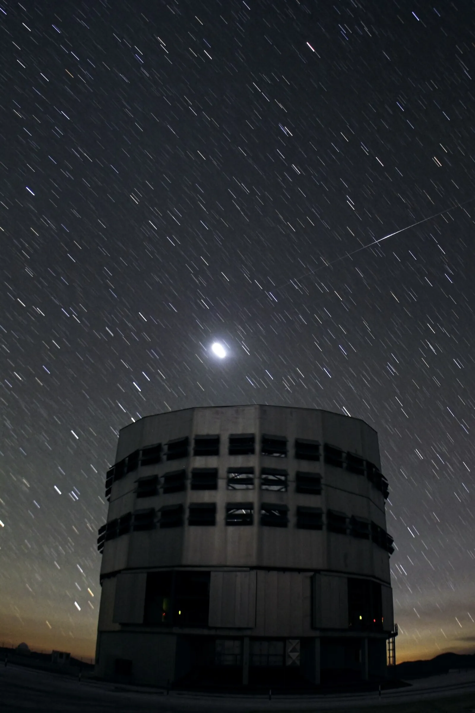 Long-exposure night photograph of a tall, cylindrical telescope building under a sky full of short star trails. A thin streak that brightens in the middle crosses the upper right, and a bright glowing spot hangs above the building.
