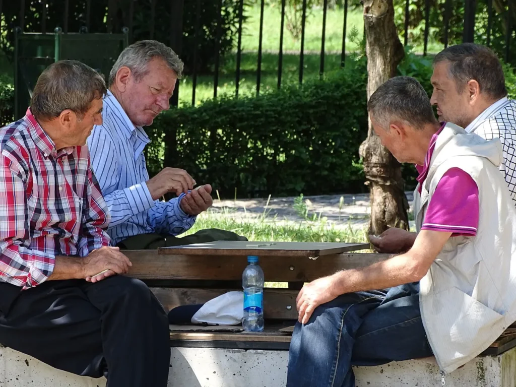 Four older men sit on a stone bench in a sunny park, leaning in over a small wooden board as they play dominoes together. A water bottle stands on the bench between them, with a hedge and iron fence behind.