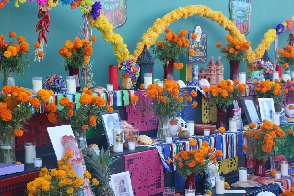 A three-tiered Day of the Dead altar covered with orange marigolds, white candles, striped cloths, paper decorations and framed photographs of loved ones.
