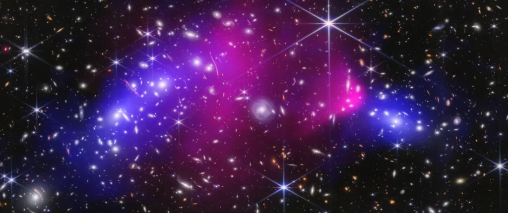 Hundreds of small galaxies and a few bright, spiky stars on a black background. A pink haze of hot gas spans the center, ending in a bright bullet-shaped patch at right, with blue haze on the left and right.