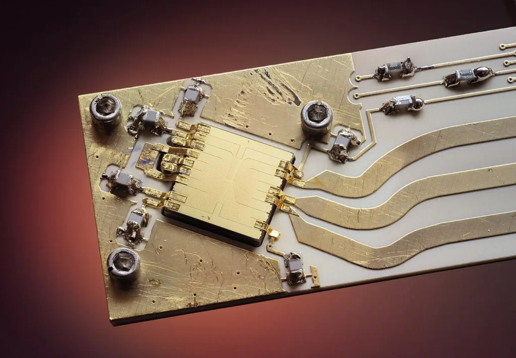 Close-up of a small square gold chip at the center of a gold-and-white circuit board, with gold wiring traces, tiny electronic components and screws, against a dark red background.