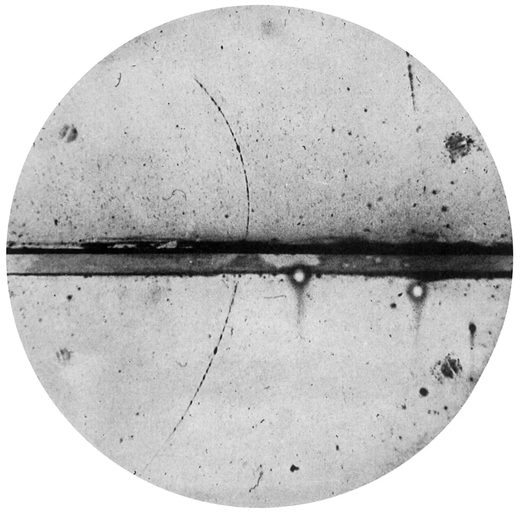 Circular black-and-white photograph crossed by a thick dark horizontal band, a lead plate. A thin dotted track rises from the lower left, passes through the plate and bends more tightly above it.