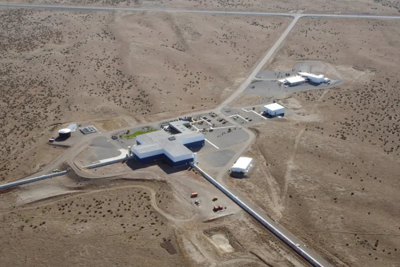 Aerial view of the LIGO Hanford detector buildings and two long perpendicular interferometer arms crossing an arid landscape.