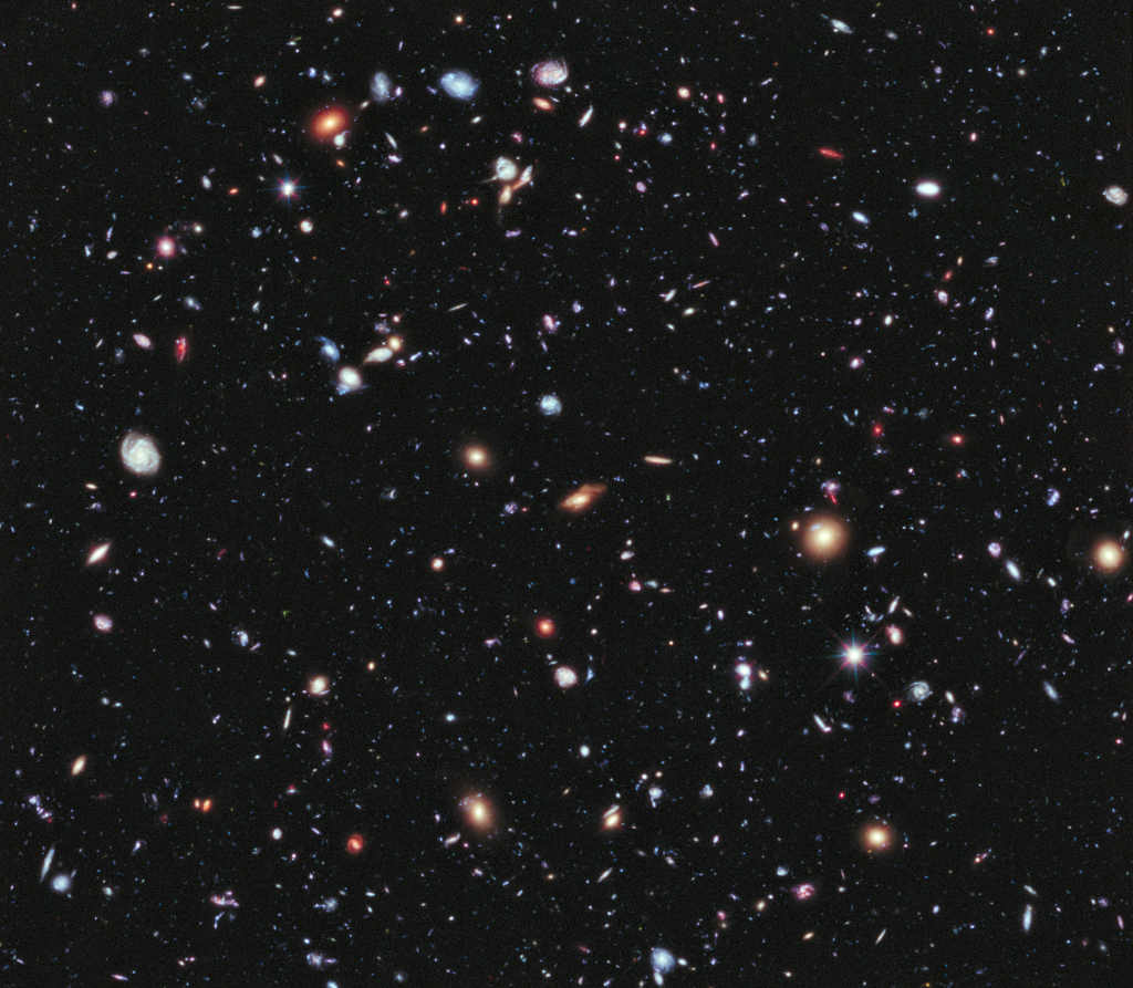 Many small blue, red, orange and white galaxies spread across a dark field, with a few bright spiked points.