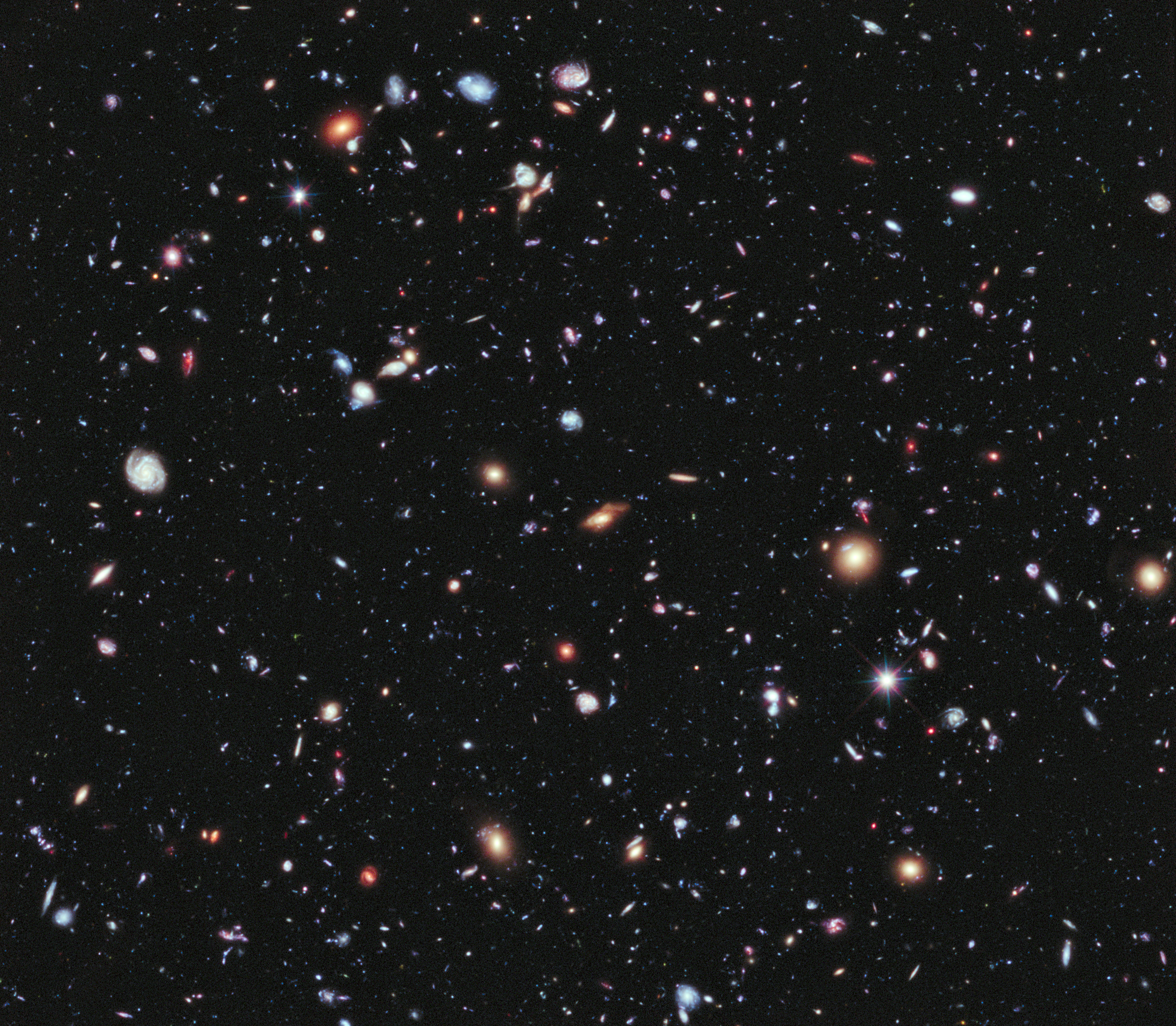 Many small blue, red, orange and white galaxies spread across a dark field, with a few bright spiked points.